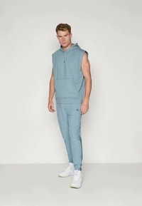 Light blue sleeveless hoodie with a front pocket, paired with matching tapered pants featuring a logo. Worn with white sneakers.
