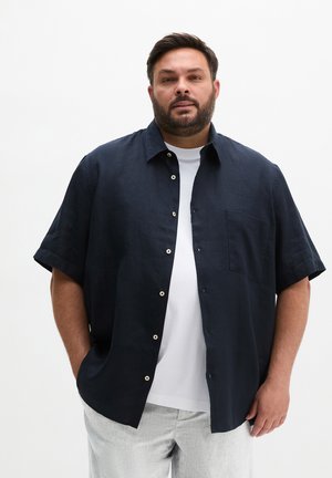 Mężczyzna w niezapiętej granatowej koszuli z krótkim rękawem, na białej koszulce i jasnoszarych spodniach na jednolitym białym tle.