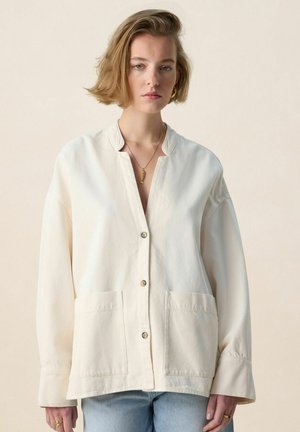 Femme aux cheveux blonds courts portant une chemise crème ample à boutons, un jean bleu clair, un collier en or et une boucle d'oreille en anneau doré, sur un fond beige.