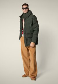 Uomo che indossa occhiali da sole neri, giacca a vento verde scuro, maglione beige, pantaloni larghi in corduroy beige e scarpe beige, in piedi su uno sfondo semplice.