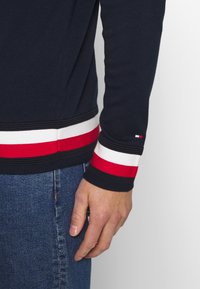 Pull en maille côtelée bleu marine avec des accents rayés rouges et blancs aux poignets et à l'ourlet. Présente une petite étiquette avec logo sur la manche. Coupe décontractée.