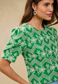 Blusa verde ricamata con maniche a sbuffo, caratterizzata da motivi floreali bianchi e dettagli in pizzo. Tessuto liscio con scollatura rotonda.