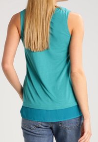Tyrkysový top bez rukávů s vrstveným lemem. Měkká látka s hladkou texturou. Bavlněná směs. Pohled zezadu ukazuje vlasy a pohodlný střih.