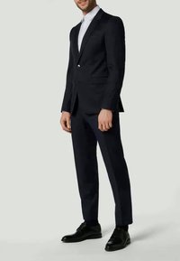 Marineblauer Anzug mit einer taillierten Jacke mit einem einzelnen Knopf und passender, maßgeschneiderter Hose. Schwarze Lederschuhe runden das Ensemble ab.