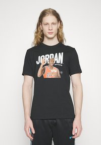 Fekete póló, grafikai nyomattal, melyen egy kosárlabdázó látható, "JORDAN" felirattal, vastag fehér betűkkel és élénk narancssárga akcentusokkal. Rövid ujjú, kerek nyakkivágású.