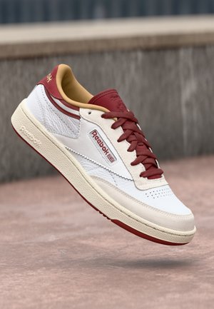 Biały i bordowy sneaker Reebok z czerwonymi sznurowadłami i beżową podeszwą, pokazany unoszący się nad betonową powierzchnią.