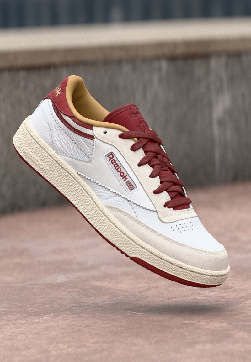 Baskets Reebok blanches et bordeaux avec des lacets rouges et une semelle beige, montrées en train de flotter au-dessus d'une surface en béton.
