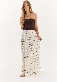 Mladá žena s dlhými blond vlasmi je oblečená v tmavohnedom topu bez ramienok a bielych sukni s vysokým pásom, na ktorých sú tmavohnedé bodky, stojí na bielom pozadí.