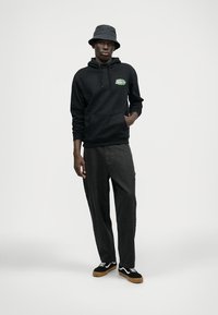 Hanorac negru cu un logo grafic verde, pantaloni gri și pantofi negri cu talpă de gumă. Modelul poartă o pălărie gri de tip bucket. Poziție în întregime.