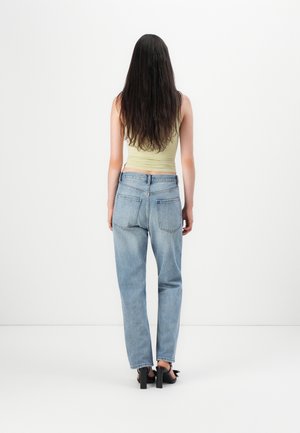 Vrouw met lang donker haar, gekleed in een lichtgroene mouwloze top, lichtblauwe jeans met hoge taille en zwarte sandalen met blokhak, staand met de rug naar de kijker.