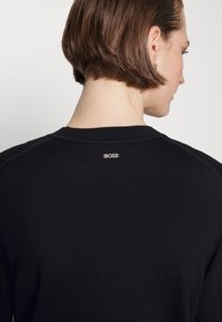 Čierny pletený sveter s okrúhlym výstrihom, na zadnej strane výstrihu s malým strieborným logom "BOSS". Mäkka textúra a priliehavý strih.