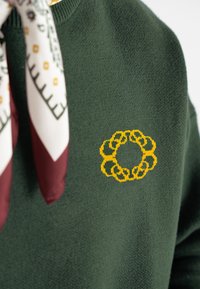 Ciemnozielony sweter o miękkiej fakturze z okrągłym, żółtym haftem kwiatowym na przodzie. Obok leży wzorzysta chusta.