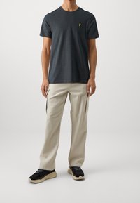 Maglietta in cotone grigia scura con colletto rotondo e piccolo logo giallo sul petto, abbinata a pantaloni cargo beige chiaro e sneaker nere.