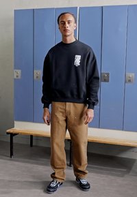 Svart hettegenser med tekstlogo, tan arbeidsbukser og svarte sneakers. Står foran blå skap med en trebenk.