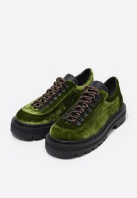 Zapatos de terciopelo verde con suelas de goma negra. Presentan un diseño de cordones con cordones marrón oscuro y acentos en el talón en contraste de color negro.