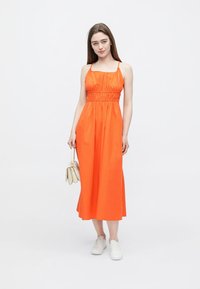 Narancssárga ujjatlan ruha megkötött derékkal, légies szoknyával és sima anyaggal. Fehér tornacipővel és világos kézitáskával párosítva.