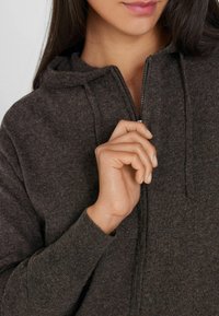 Sudadera con cremallera en color marrón, de textura suave, que incluye capucha, cremallera frontal y puños acanalados. Mano tirando suavemente de la cremallera.