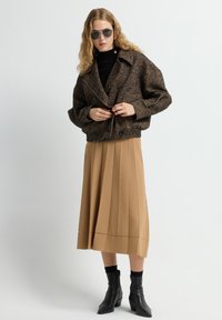 Bruine oversized jas met een visgraatpatroon, zwarte coltrui, en beige geplooide midi-rok, gecombineerd met zwarte enkellaarsjes en een zonnebril.