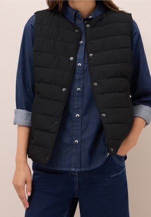 Femme portant un gilet matelassé noir par-dessus une chemise en denim bleu aux manches retroussées et un jean bleu foncé, une main dans la poche.