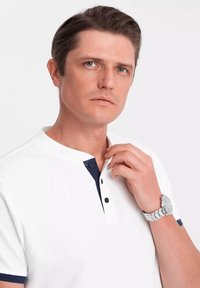 Bílá polo košile s námořnickým límcem a dvěma černými knoflíky. Model má na sobě stříbrné hodinky, krátké vlasy a neutrální výraz.