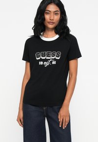 Žena s dlouhými černými vlasy, oblečená v černém tričku s bílým límečkem a textem "GUESS 1981 est.", v kombinaci s tmavě modrými džíny, stojící na bílém pozadí.