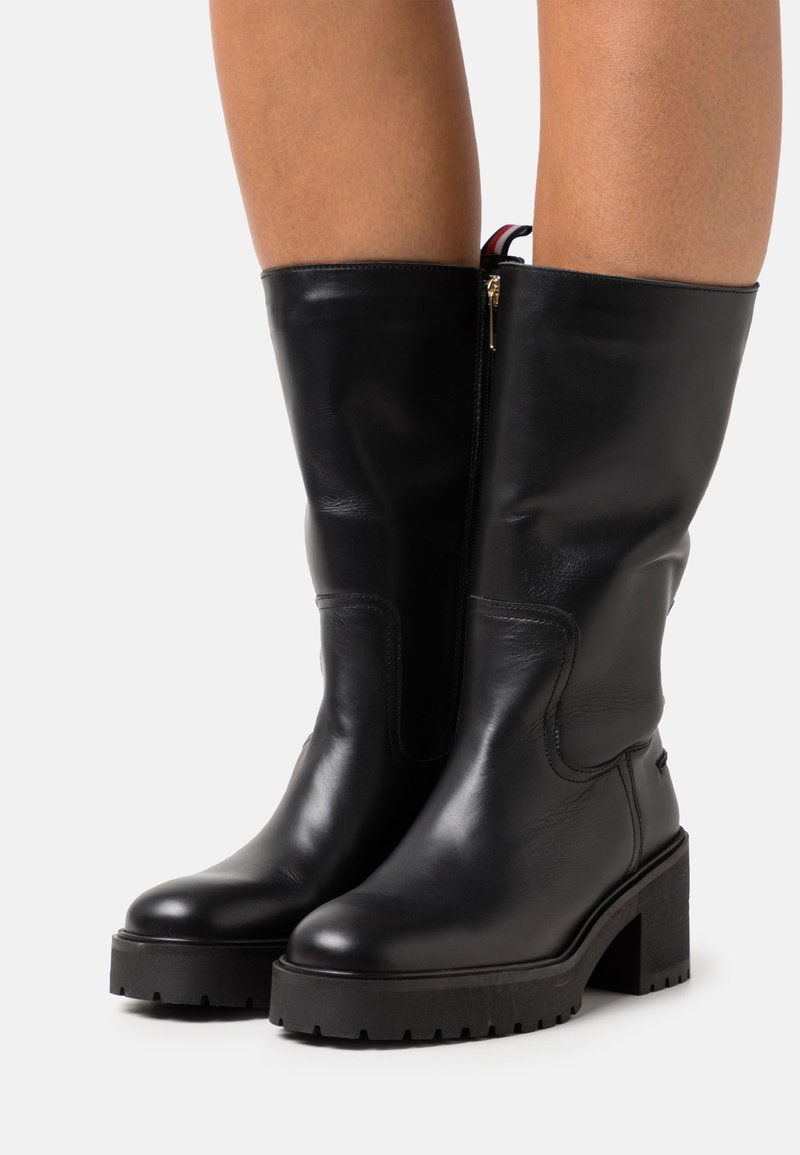 Bottes noires en cuir, montant jusqu'au genou, avec un talon épais et une surface texturée. Présente une fermeture éclair latérale et un accent contrastant en haut.