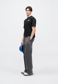 Czarny t-shirt z krótkim rękawem, szare spodnie dresowe, szare tenisówki. Trzyma niebieską czapkę. Na koszulce i spodniach logo, luźny krój.