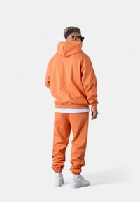Haut à capuche orange et joggers assortis, avec des poignets côtelés, une texture douce, des accents blancs au niveau de l'ourlet, associés à des baskets blanches.