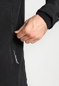 Veste softshell noire avec une poche zippée, arborant le logo "ClimaProtect". Le tissu est lisse, extensible et conçu pour une utilisation en extérieur.