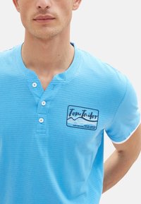 Svetlomodrá polokošeľa s krátkym rukávom z textúrovanej látky, s henley výstrihom, nášivkou s logom "Tom Tailor" a tromi gombíkmi na golieri.