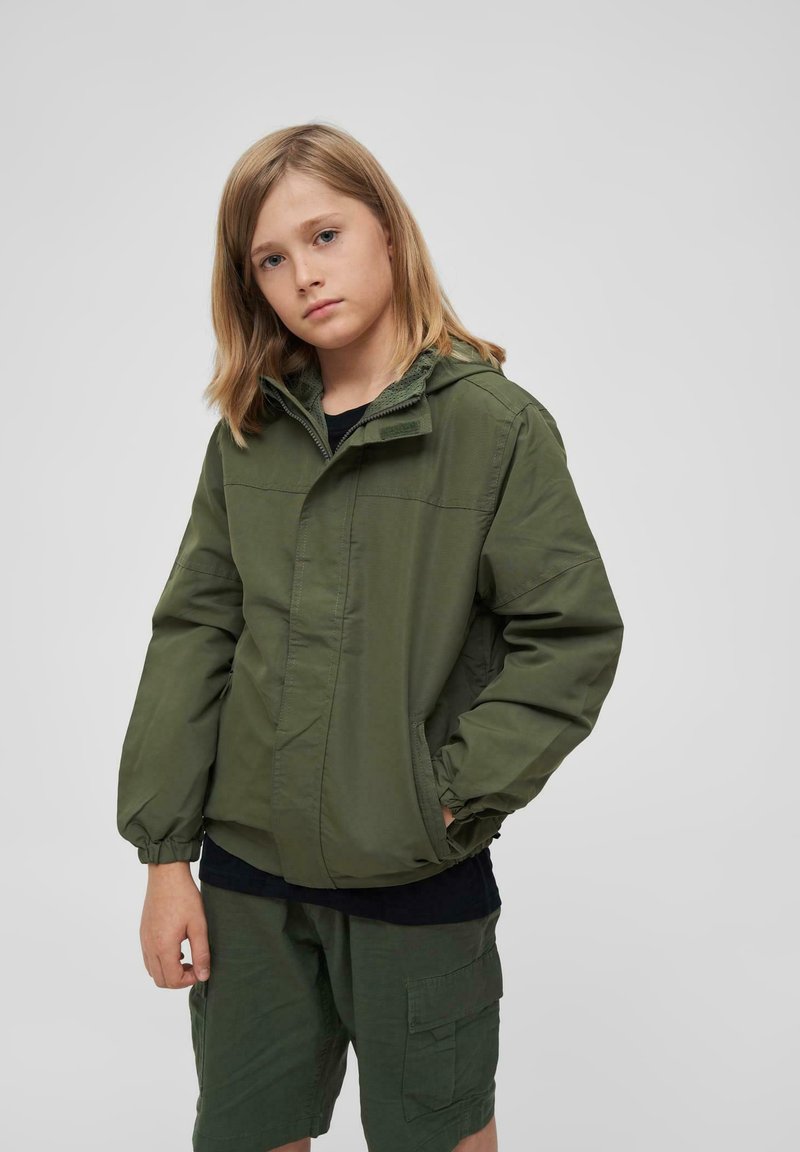 Veste légère verte avec capuche, poignets élastiques et poches avant. Portée sur une chemise noire et associée avec un short cargo vert.