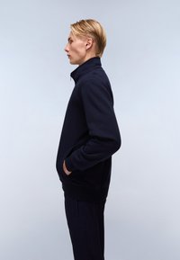 Marineblå fleecejakke med høy krage, glidelås og sidelommer. Har ribbestrikkede mansjetter og kant. Glatt tekstur og enkel design.