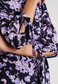 Zbliżenie na ramię kobiety w kwiecistej bluzce z fioletowymi i różowymi wzorami na czarnym tle, zawiązanej w kokardę przy mankiecie.