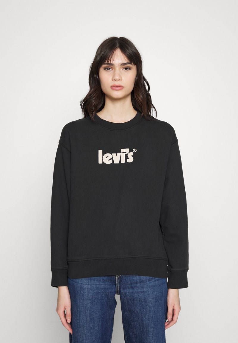 Sweatshirt noir en coton, arborant un grand logo beige 'Levi's' sur la poitrine, avec des poignets et un ourlet côtelés. Porté avec un jean bleu.