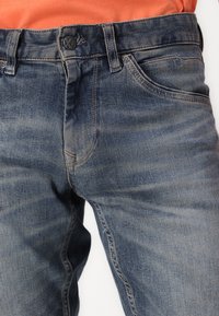 Jeans de mezclilla azul oscuro con un aspecto desgastado, que cuentan con un diseño clásico de cinco bolsillos y un cierre de botón en la cintura.