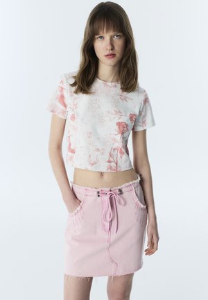 Joven mujer con cabello castaño liso, llevando un crop top blanco con flores y una falda rosa claro con cordón y bolsillos delanteros, de pie con las manos en los bolsillos.