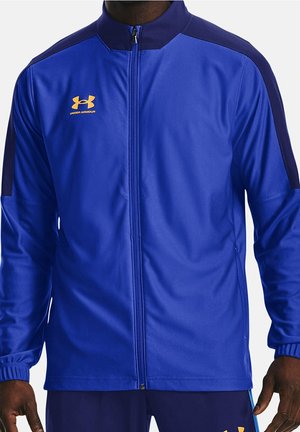 Mężczyzna ubrany w niebieską kurtkę Under Armour na zamek z ciemnoniebieskimi paskami na ramionach oraz pasujące niebieskie spodnie.