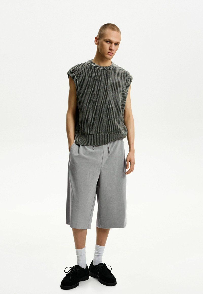 Top in maglia grigia a maniche corte con trama, pantaloni corti grigi larghi con coulisse, calze bianche alla caviglia, scarpe in suede nere, design minimalista contro uno sfondo neutro.