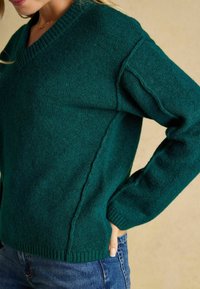 Pulover tricotat texturat, verde, cu decolteu în V, tiv și mansete ribbate. Prezintă detalii pe cusăturile laterale și are o croială lejeră, asortat cu blugi.