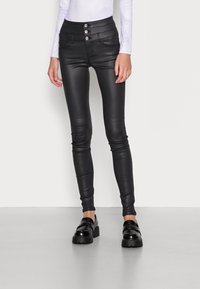 Pantalon noir en similicuir taille haute avec fermeture à trois boutons, coupe slim et texture lisse, associé à des chaussures à plateforme noires épaisses.