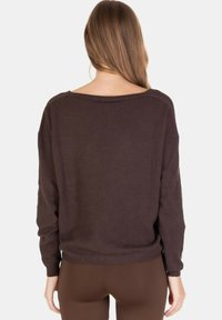 Donna con capelli lunghi castano chiaro che indossa un maglione marrone scuro ampio e pantaloni marroni aderenti, vista da dietro su uno sfondo bianco.