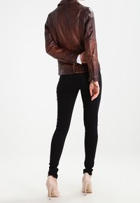 Giacca in pelle marrone con silhouette aderente, jeans skinny neri e décolleté nude. La giacca presenta polsini con zip e un colletto alto.