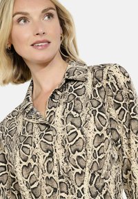 Žena s krátkými blond vlasy, oblečená v košili na knoflíky s bežovým a černým hadím vzorem, dívá se mírně nahoru a na stranu.