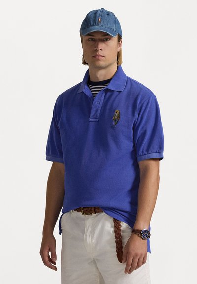 Cămașă polo albastră cu guler și logo brodat, purtată cu o cămașă cu dungi dedesubt. Combinată cu pantaloni albi și o curea împletită maro.