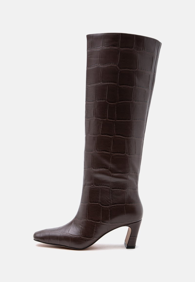 Botte haute en cuir marron foncé jusqu'au genou avec texture crocodile, bout pointu et talon bas incurvé sur fond blanc.