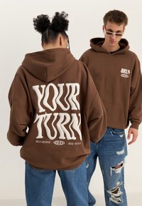 Brun oversized hette med hvit hevet tekst "YOUR TURN" på ryggen og logo på fronten, kombinert med slitte blå jeans.