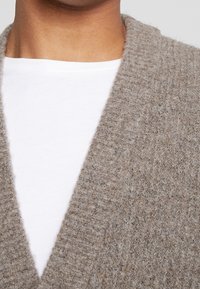 Beige stickad kavaj med en strukturerad yta, med en djup V-ringning och synliga ribbade kanter, lagd över en vit skjorta.