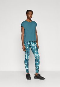 Επάνω μέρος κοντομάνικο σε πετρόλ απόχρωση συνδυασμένο με πετρόλ κολάν που φέρει γεωμετρικό λευκό σχέδιο, όλα συμπληρωμένα με μαύρα αθλητικά παπούτσια.