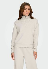 Lys beige pullover med høj krave, lynlås-lukning, afslappet pasform og lange ærmer. Glat tekstur med ribbede manchetter og forneden.