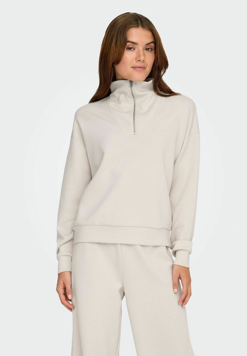 Lys beige pullover med høj krave, lynlås-lukning, afslappet pasform og lange ærmer. Glat tekstur med ribbede manchetter og forneden.